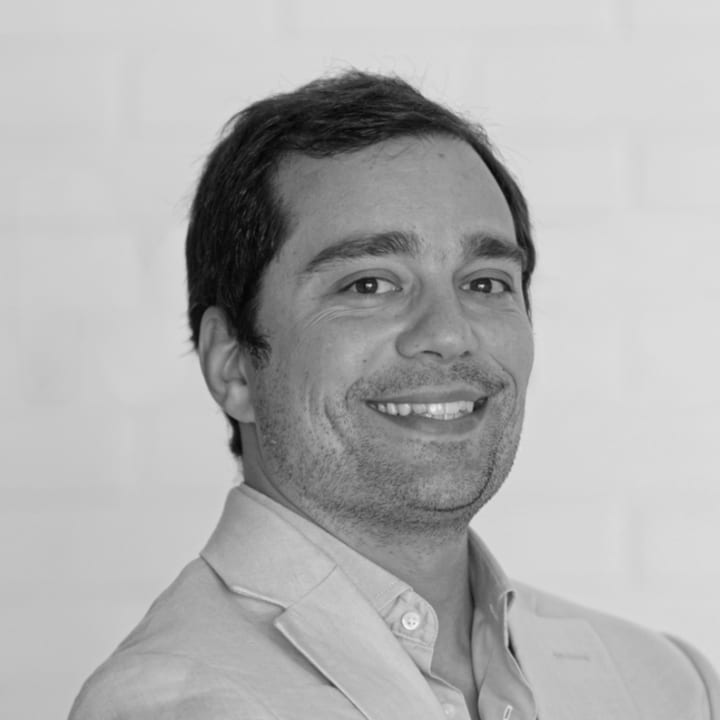 Manuel Faria, do peito para cima, o corpo ligeiramente de lado e o rosto virado para a câmara, com um sorriso aberto. Barba curta, casaco de linho sobre camisa de colarinho aberto. Fundo liso e claro, sem outros elementos. A preto e branco.
